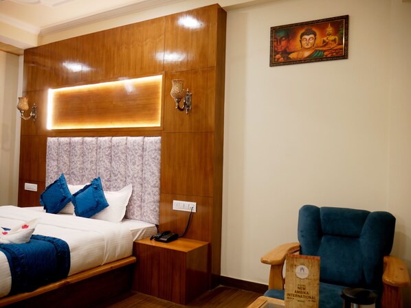 Una habitación de hotel con cabecera de madera, una cama con ropa de cama azul y blanca, una mesita de noche con una lámpara, un sillón azul y un cuadro de Buda enmarcado en la pared.