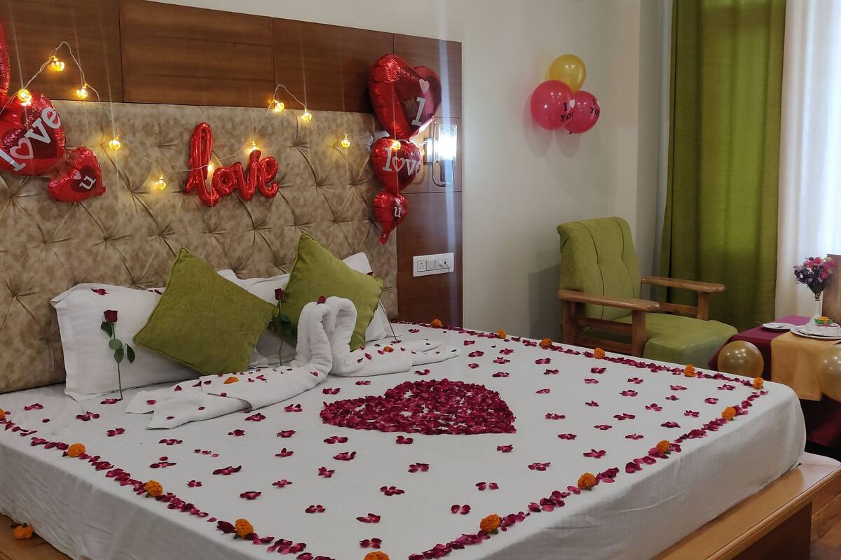 A bedroom with a tufted headboard, a bed with rose petals and a heart-shaped decoration, a green armchair, and a table with a bouquet of flowers.