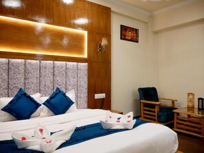 Habitación de hotel con una cama grande, almohadas azules y blancas, cabecera de madera, una mesita con lámpara y una silla.
