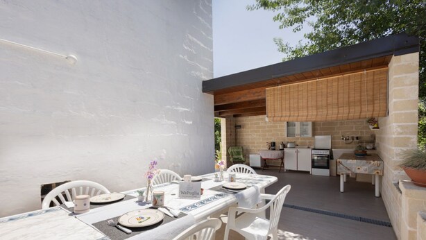 Un área de comedor al aire libre con una mesa para dos personas, una cocina al fondo y una pared de piedra.