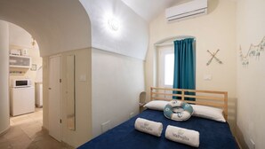 Une chambre avec un lit en bois, une fenêtre avec des rideaux bleus, un appareil de climatisation et, au fond, une petite kitchenette.
