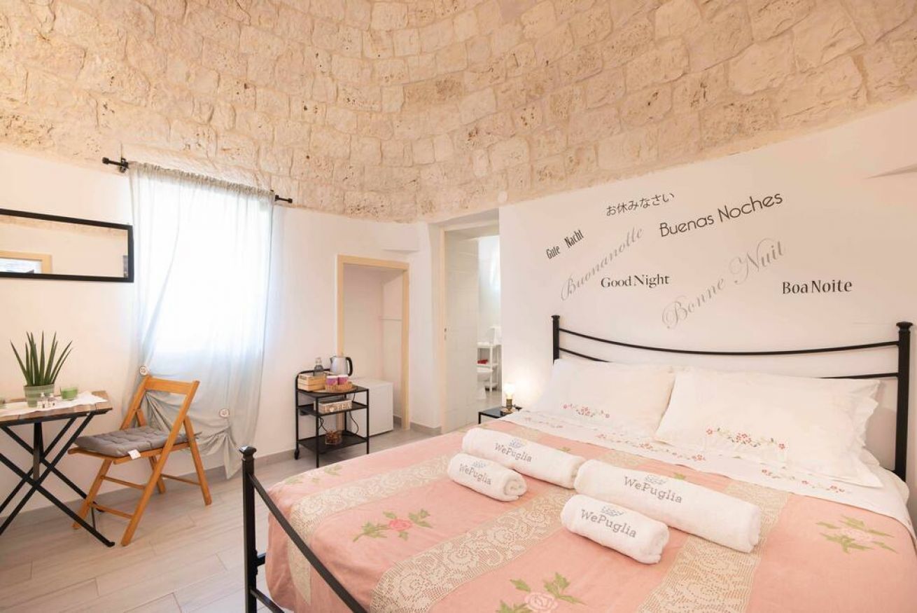 A cozy bedroom with a stone wall, a bed with floral bedding, a desk with a chair, and a nightstand with a lamp.