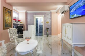A room with a marble floor, a white table with a glass top, a white cabinet with gold accents, a television mounted on the wall, and a door with a sign indicating the room. - Via Villa Iliotti (Rhodes)