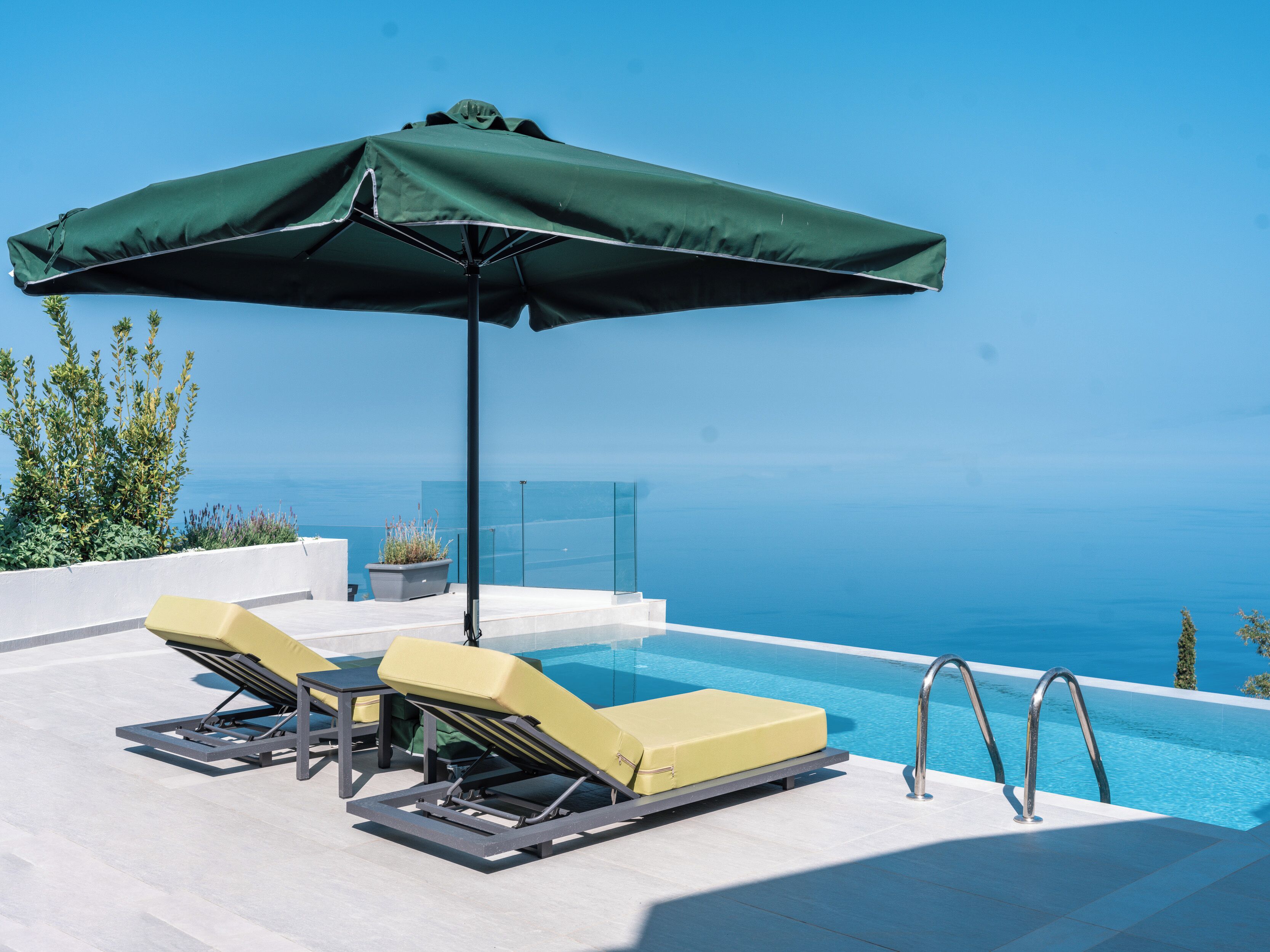 A poolside area with two sun loungers and a green umbrella, overlooking a clear blue sea.