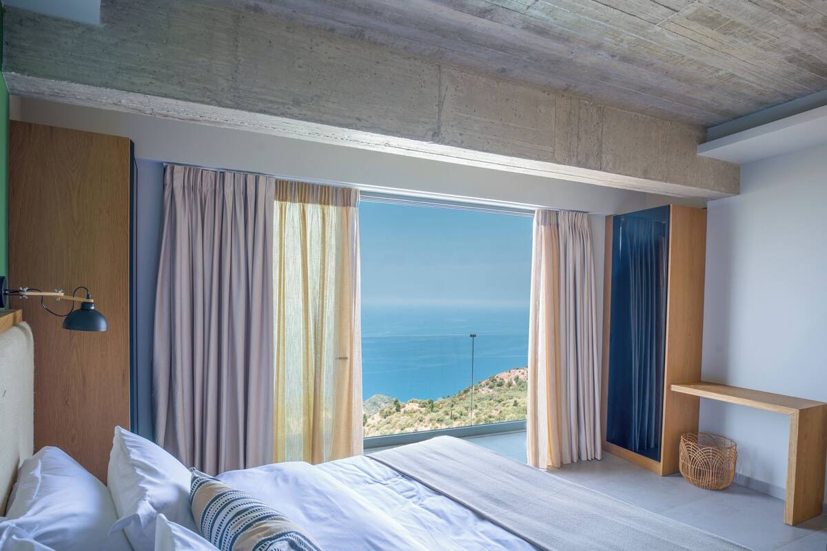 A bedroom with a large bed, a wooden headboard, a bedside lamp, and a view of the sea.