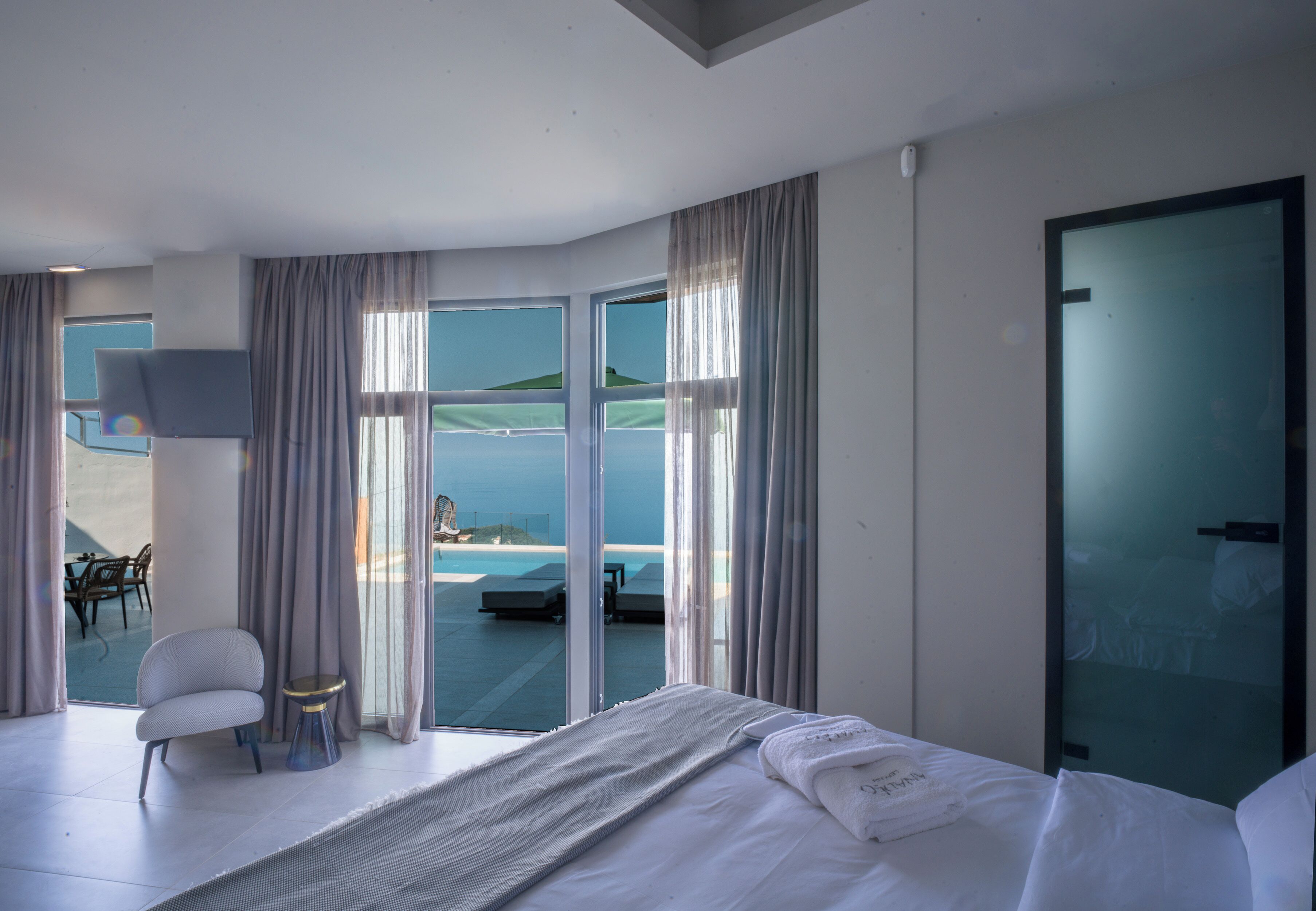 A modern hotel room with a large bed, a small round table, a chair, and a view of a pool and beach.