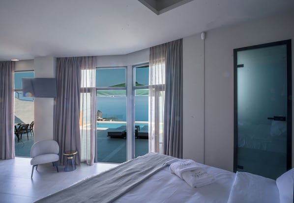 Habitación de hotel moderna con una cama grande, una mesita redonda pequeña, una silla y vistas a una piscina y playa.