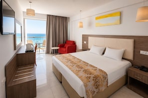 Habitación de hotel con cama, mesita de noche, escritorio con silla, sillón rojo y vista al mar.