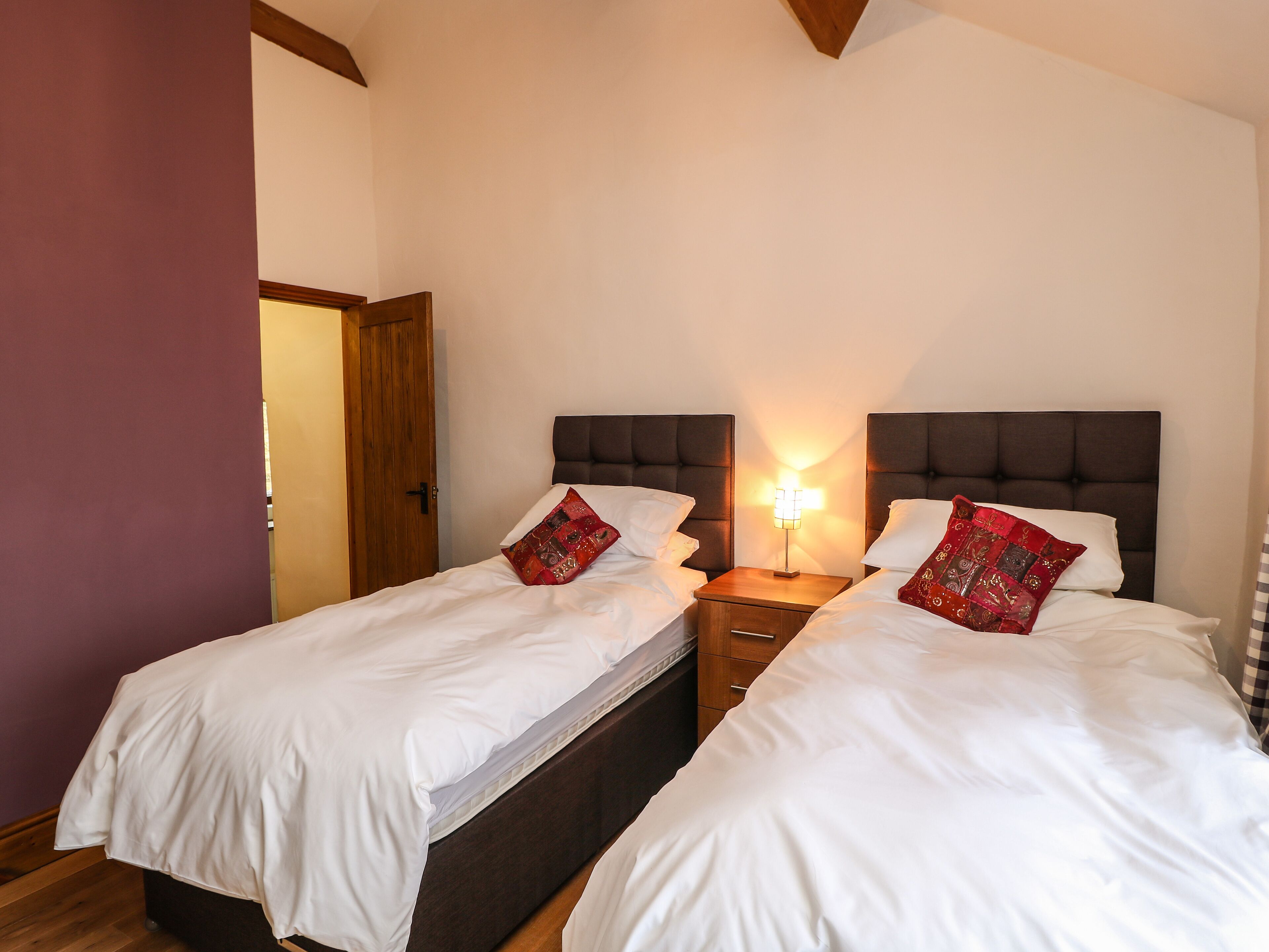 A double room with two beds, each with white bedding and red decorative pillows. There is a wooden nightstand with a lamp between the beds.