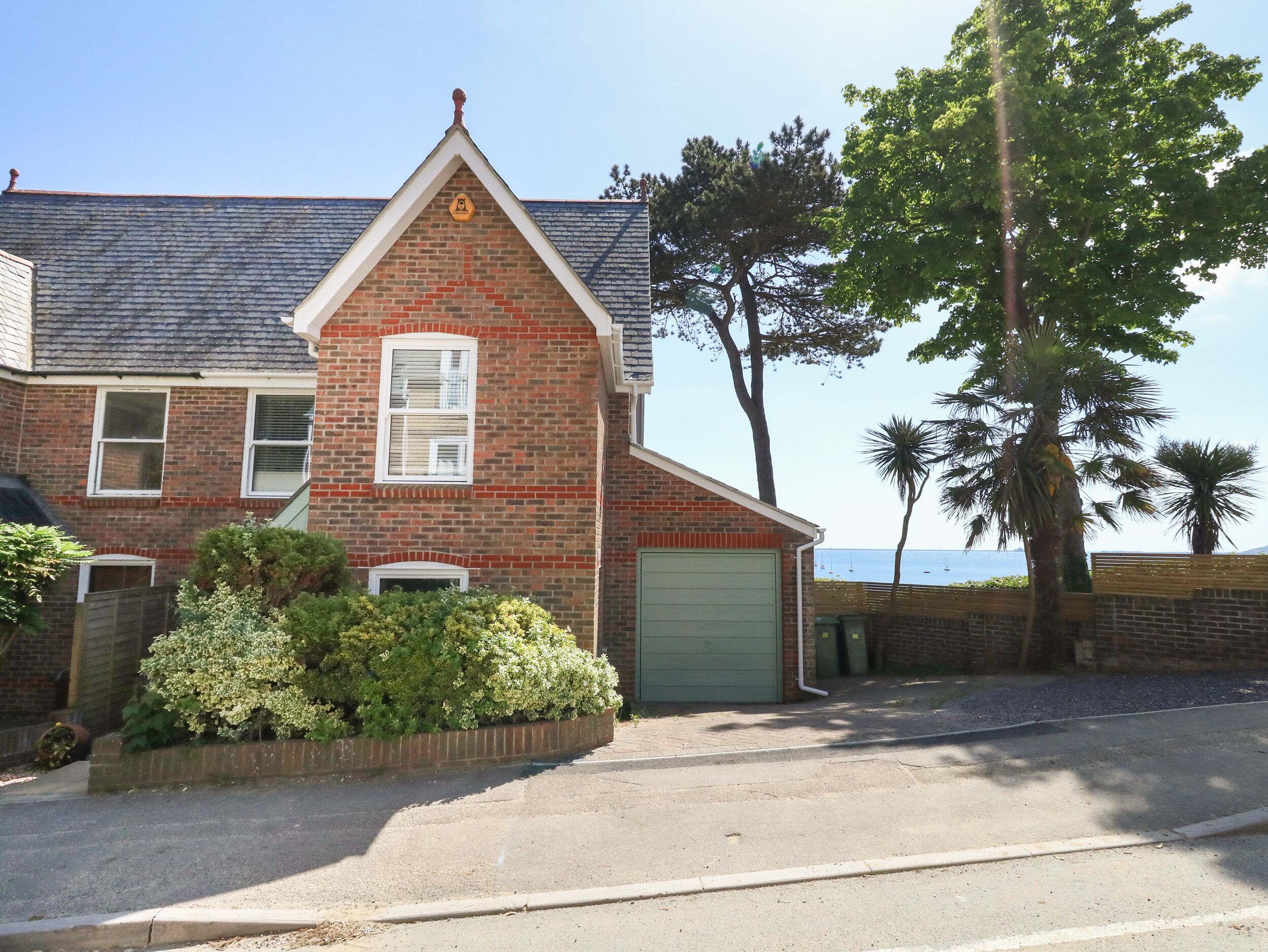 A two-story brick house with a green garage door, a small front garden, and a view of the sea.