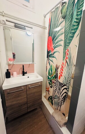 A bathroom with a white sink, wooden cabinet, mirror, and a shower curtain with a tropical leaf pattern. - Appart "L'échappée Belle" Montpellier center, Train station (Montpellier)