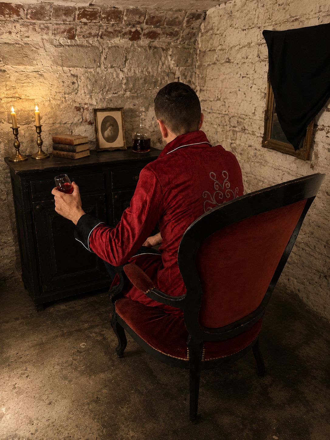 Une personne est assise sur une chaise dont le dossier est recouvert de velours rouge et tient un verre de vin rouge. En arrière-plan, on aperçoit un meuble en bois sombre, orné d’un tableau encadré et d’un chandelier avec deux bougies allumées.
