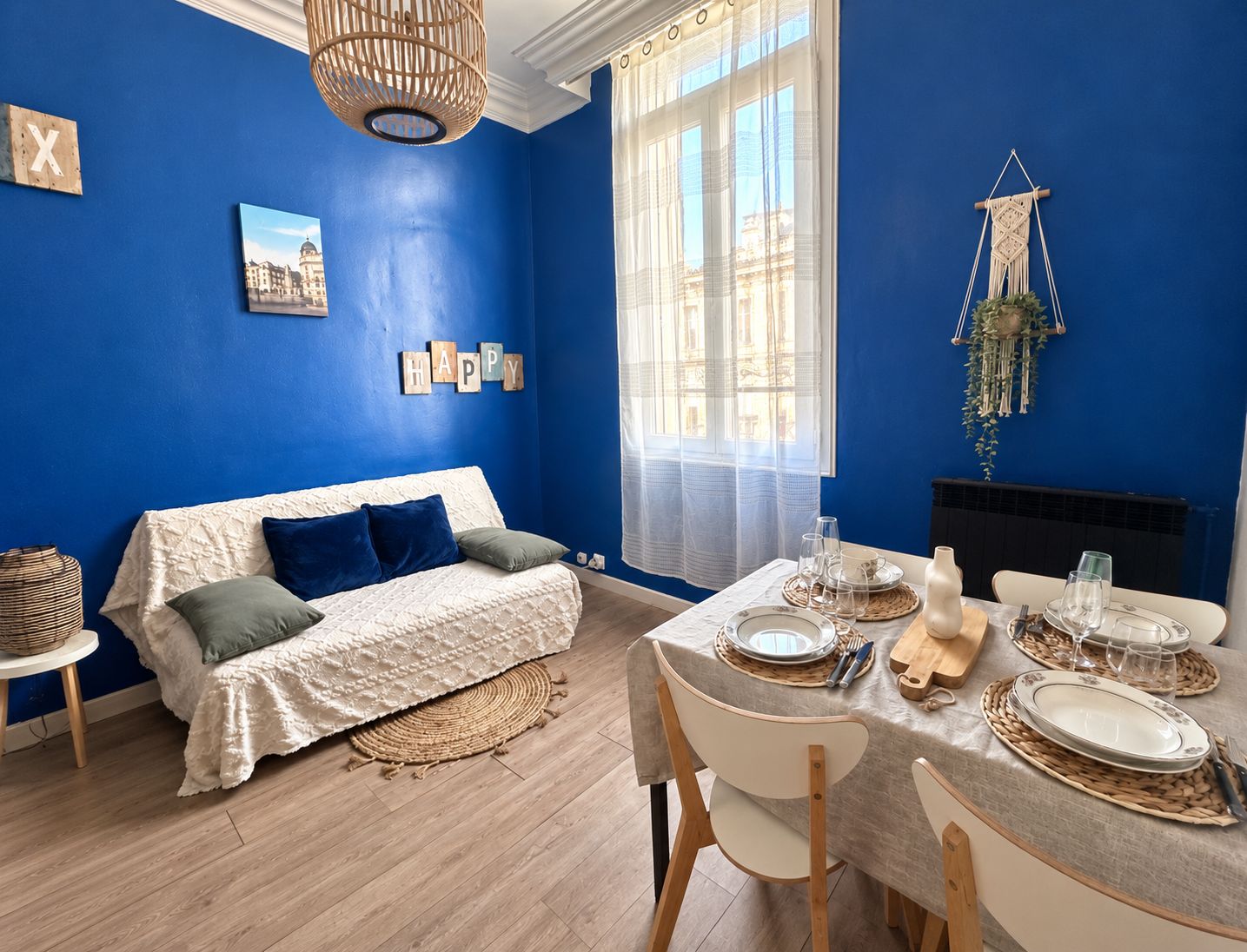 A room with a blue wall, a white sofa, a dining table set with plates and glasses, and a hanging macrame plant.