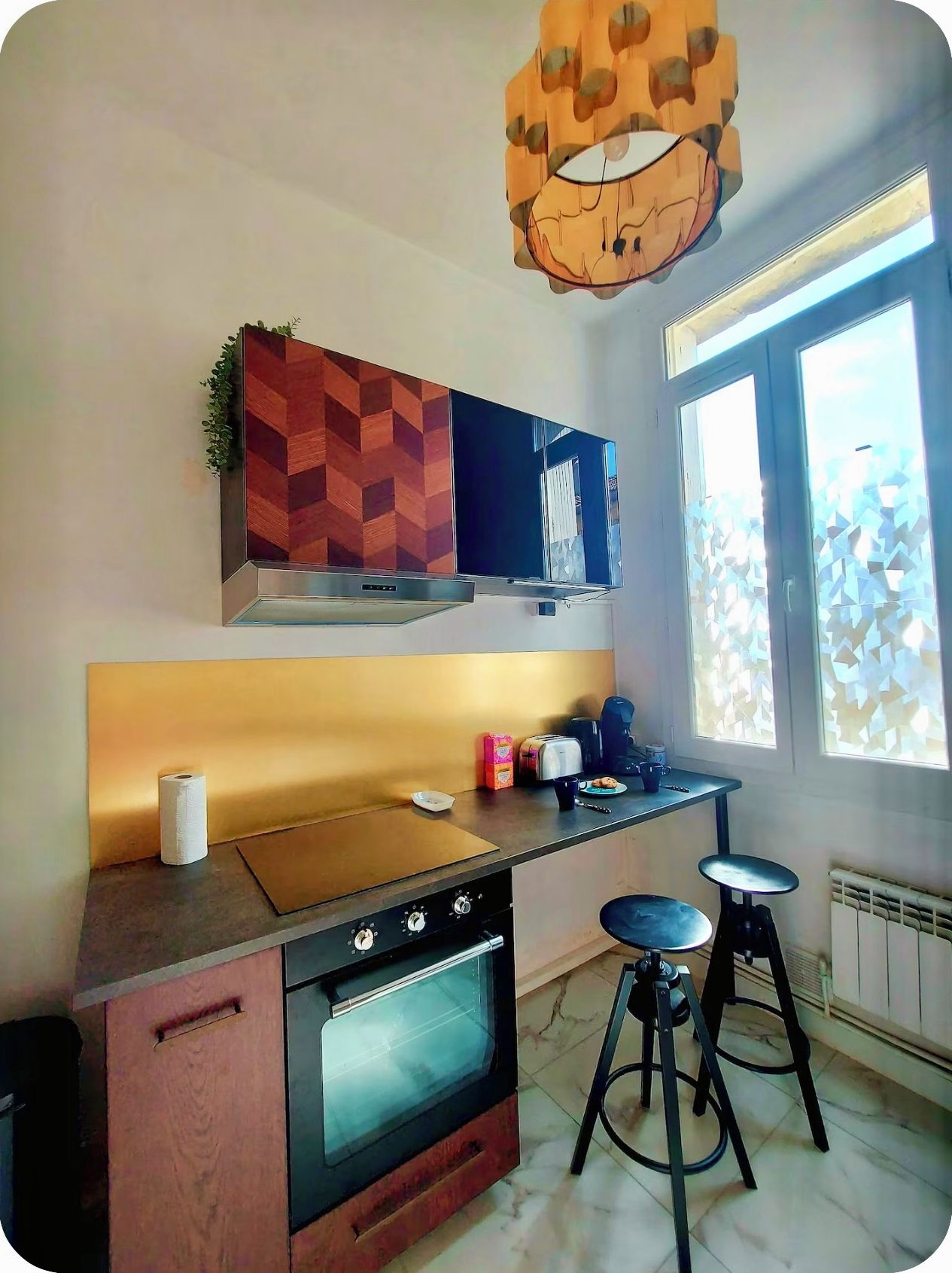 Une cuisine équipée d’un four encastré, d’une cuisinière à gaz, d’un four à micro-ondes, d’une machine à café et d’une fenêtre avec des stores à motifs.