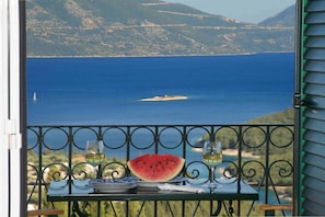 Une table avec des assiettes, un verre de vin et une tranche de pastĂšque, le tout placĂ© sur un balcon donnant sur une Ă©tendue dâeau et sur une terre lointaine.