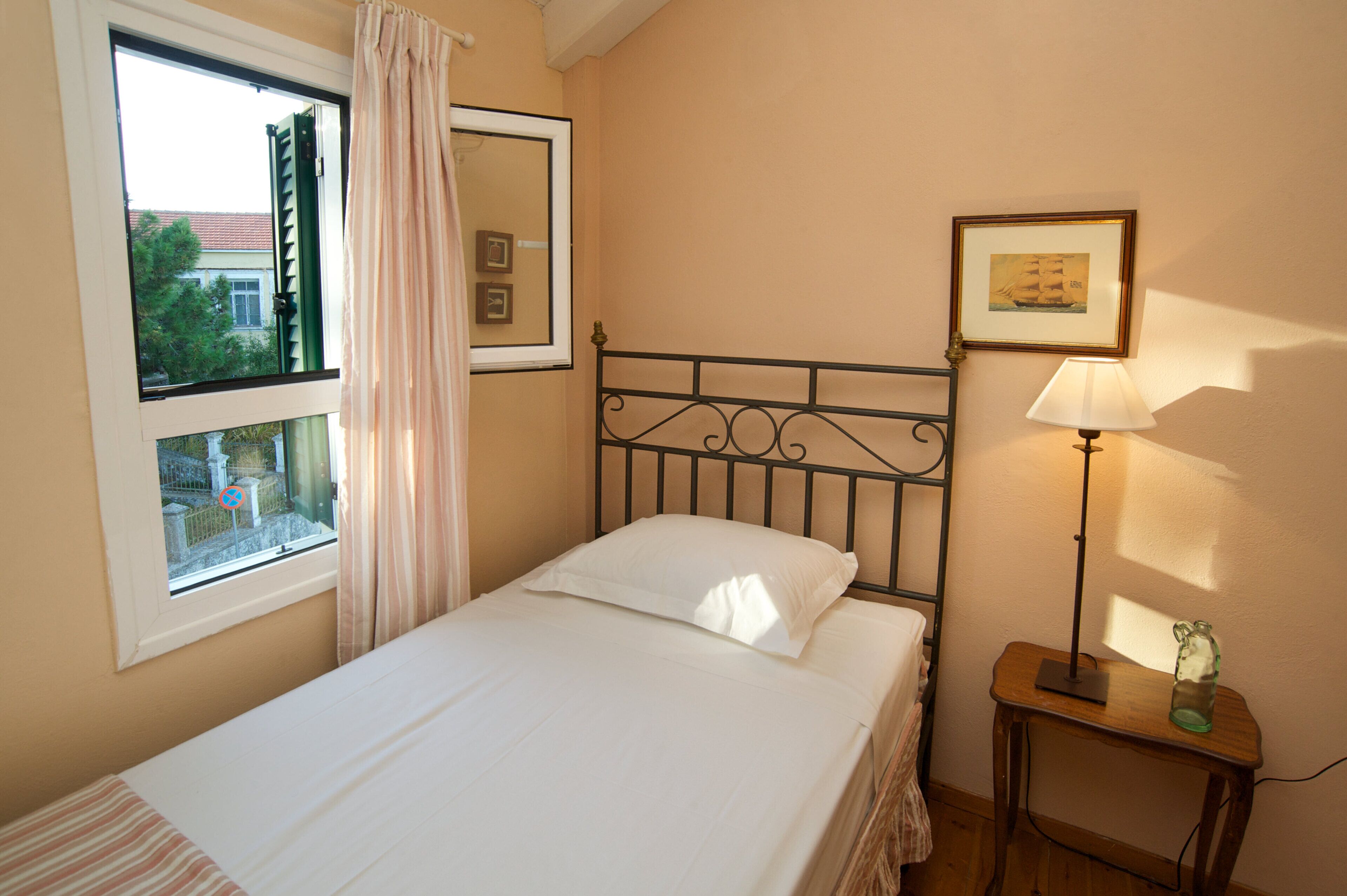 Una cama individual con cabecera de metal, una mesita de noche con lámpara, una ventana con vista a árboles y una casa, y un cuadro en la pared.
