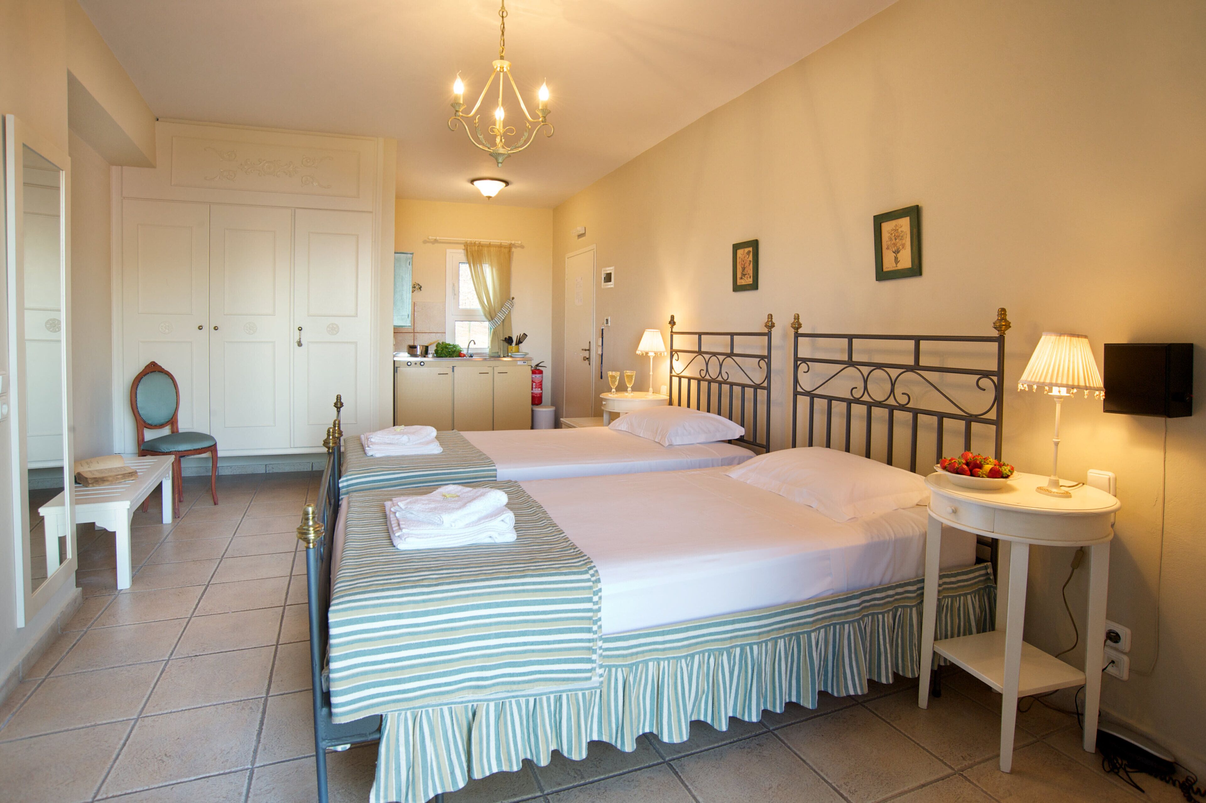 A hotel room with two beds, a sitting area with a chair, a small table with a lamp, and a kitchenette in the background.