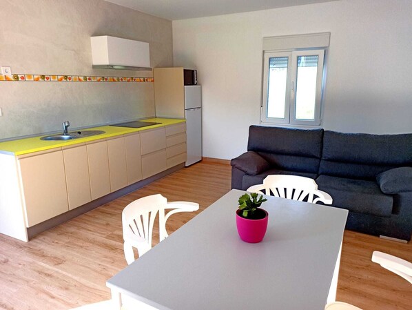 A compact kitchen with a sink, a microwave, and a refrigerator. There is a dining table with chairs and a potted plant. A dark sofa and a window with blinds are also visible.