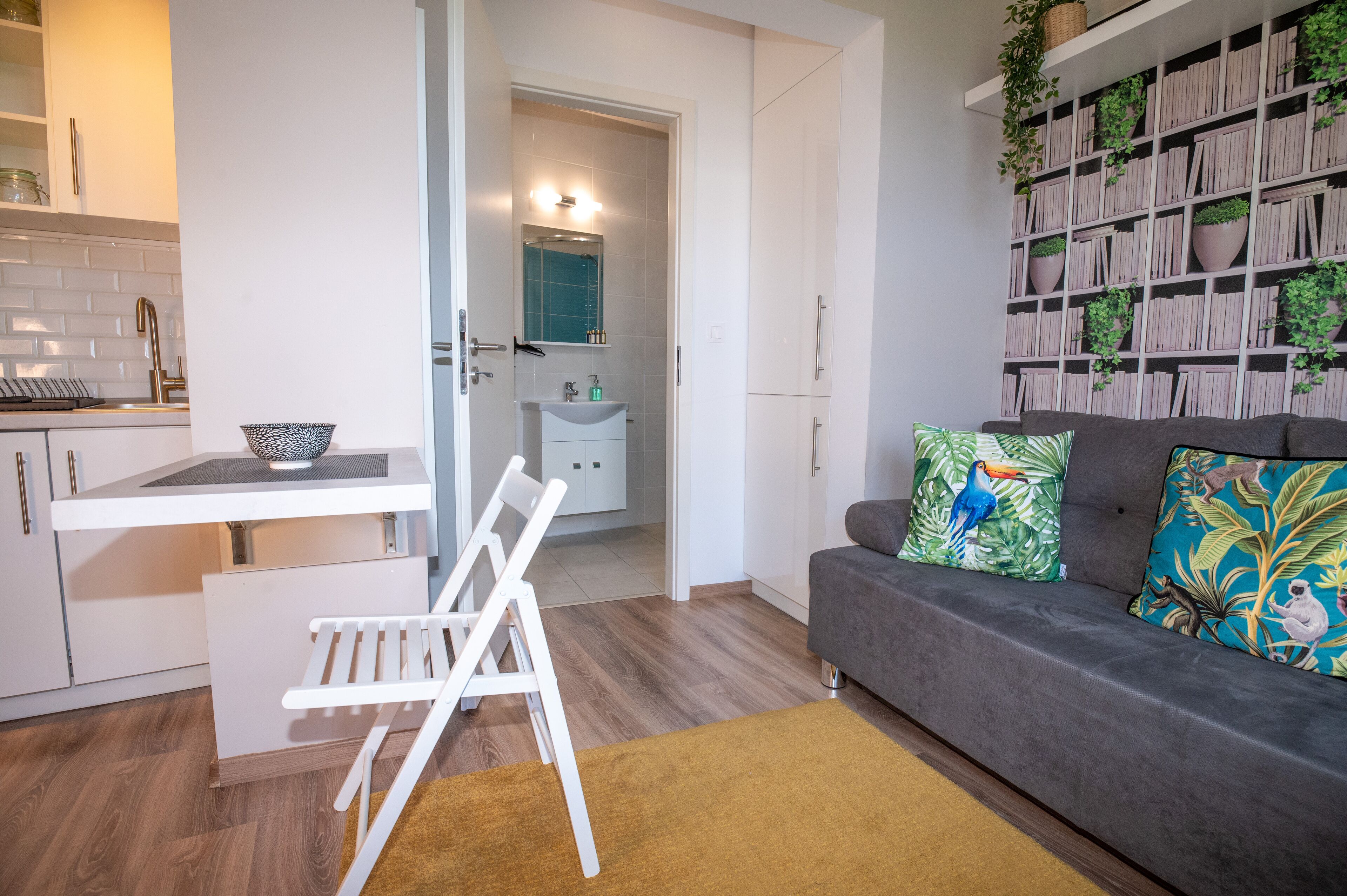 A modern living room with a grey sofa, colorful pillows, a white chair, and a bookshelf filled with books and plants.