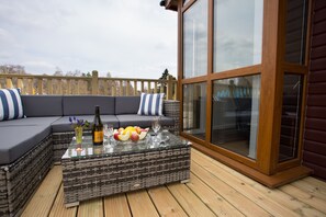 A wicker sofa with cushions, a glass coffee table with fruits and a bottle, and a wooden deck.