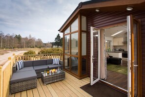 Eine Holzterrasse mit einer Couch und einem Esstisch, eine Glastür, die in eine Küche führt, und Blick auf eine natürliche Landschaft.