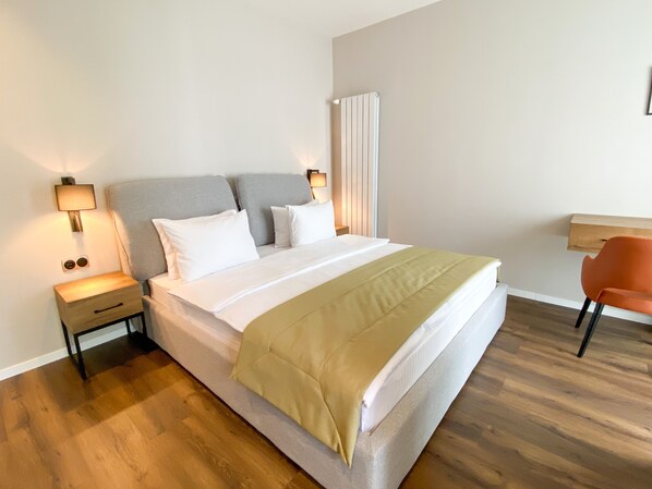 Un dormitorio ordenado con cama, mesitas de noche, una silla y un radiador.