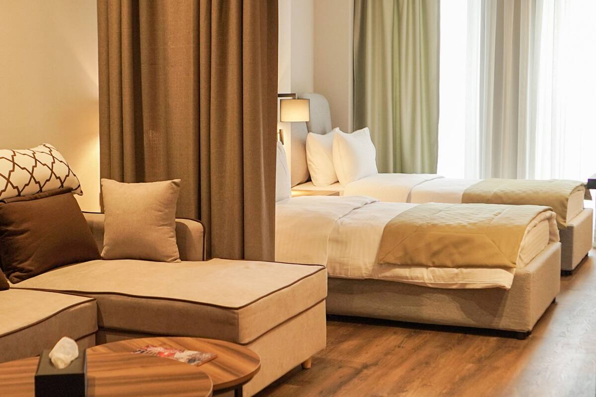 A hotel room with a sofa, two beds, a bedside table, and a lamp.