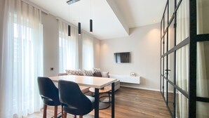 Un soggiorno moderno con zona pranzo, caratterizzato da un'ampia finestra con tende leggere, un divano bianco, una TV a parete e un pavimento in legno.