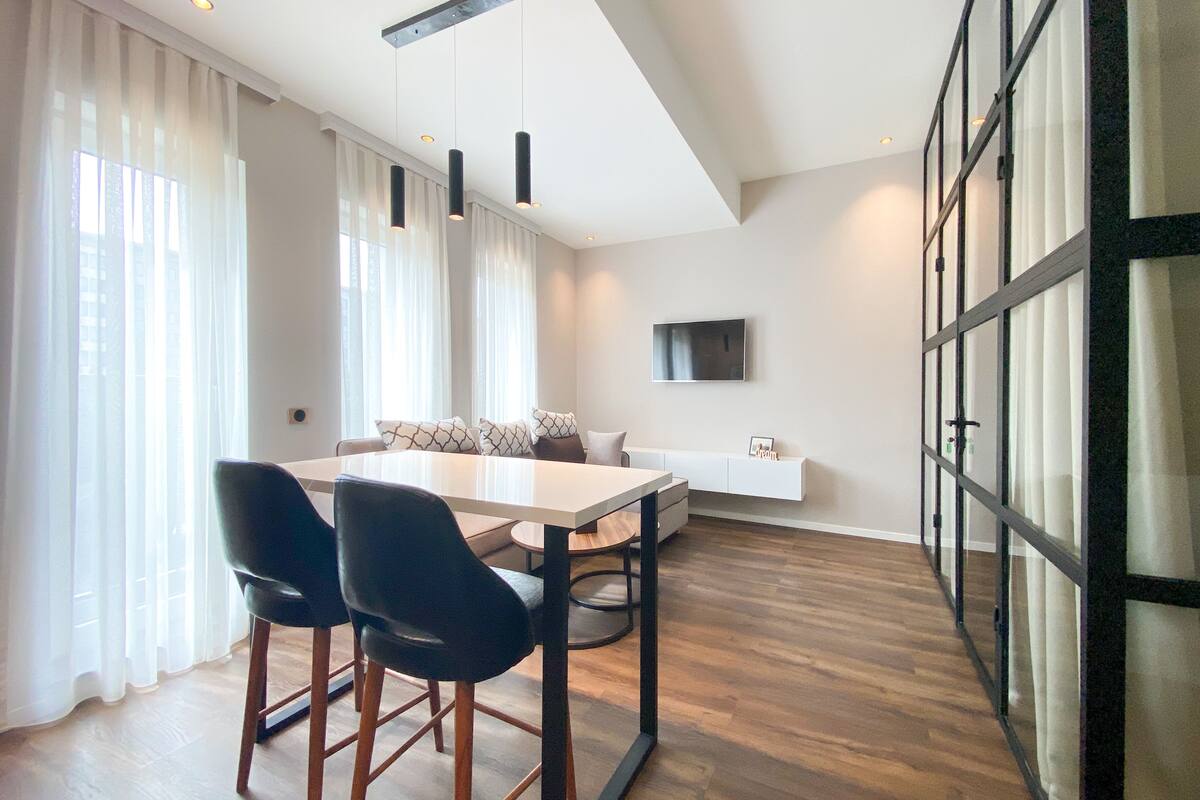 A modern living room with a dining area, featuring a large window with sheer curtains, a white sofa, a TV mounted on the wall, and a wooden floor.