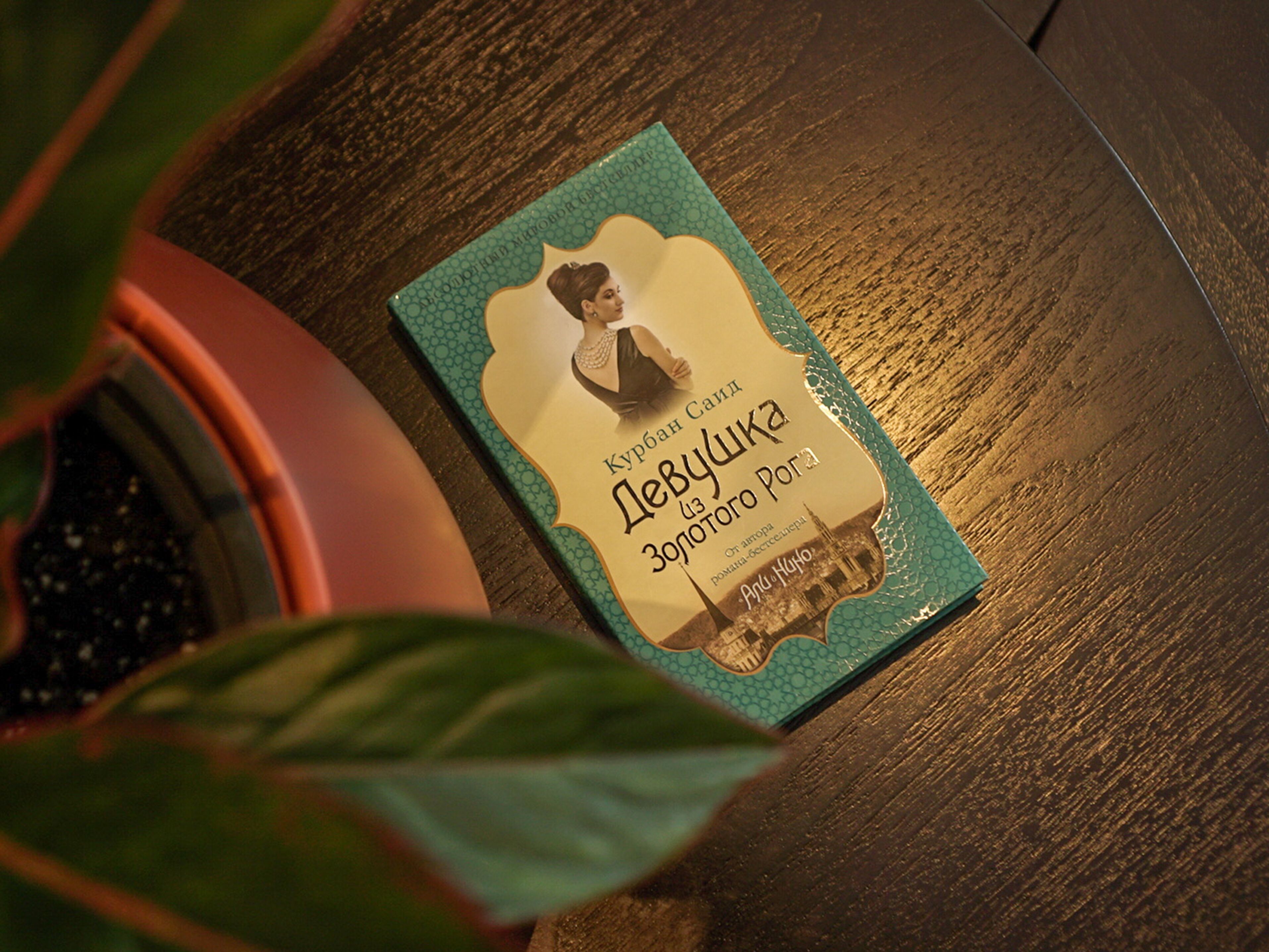 A book titled 'Курбан Саид Дебушка' with a cityscape illustration on the cover, placed on a wooden surface next to a potted plant.