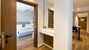 Una camera d'albergo con pavimento in legno, un letto con lenzuola bianche, un comodino con una lampada e uno specchio grande.