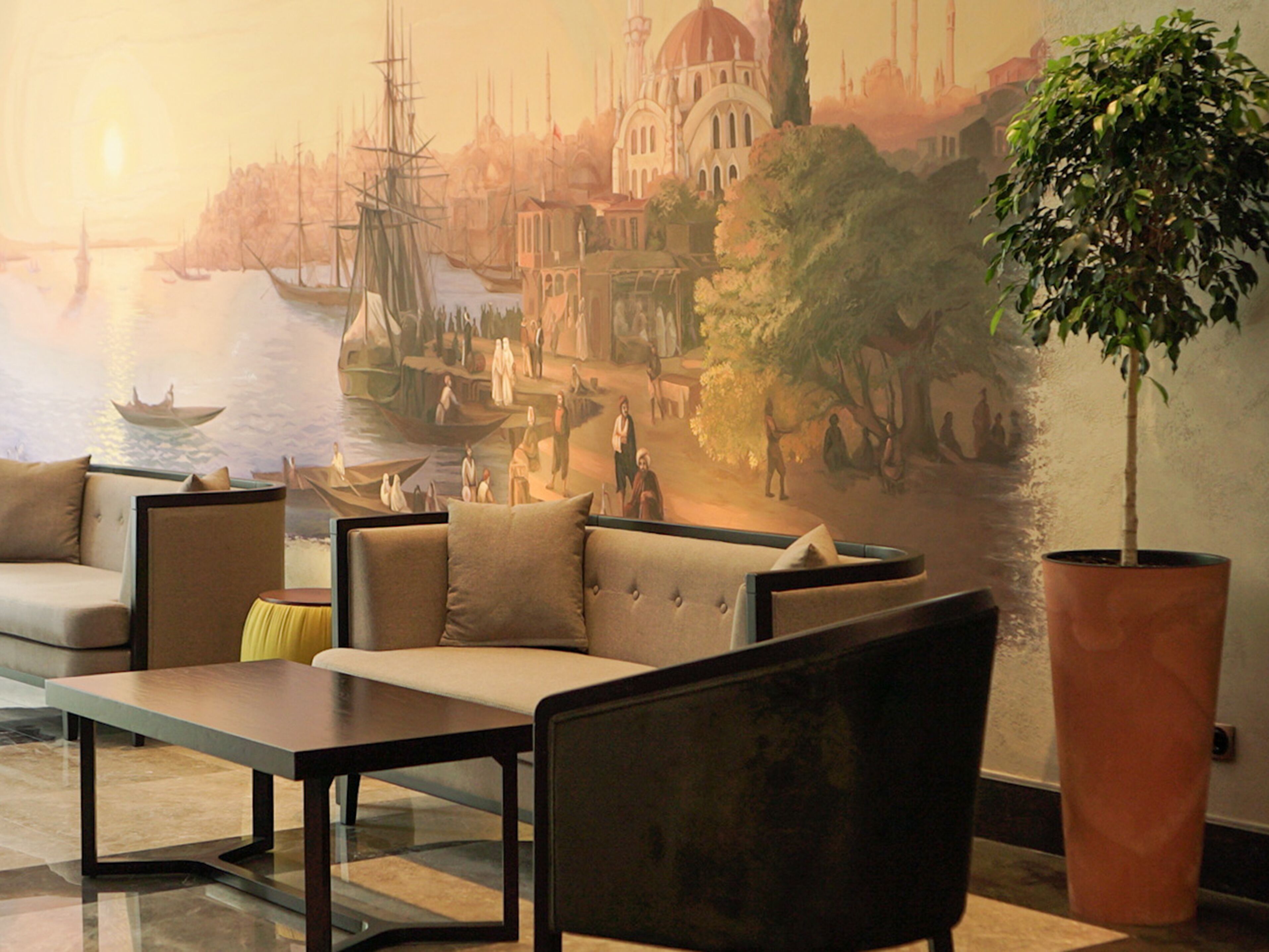 A modern lounge with a mural of a historical cityscape, seating arrangements, and a large potted plant.