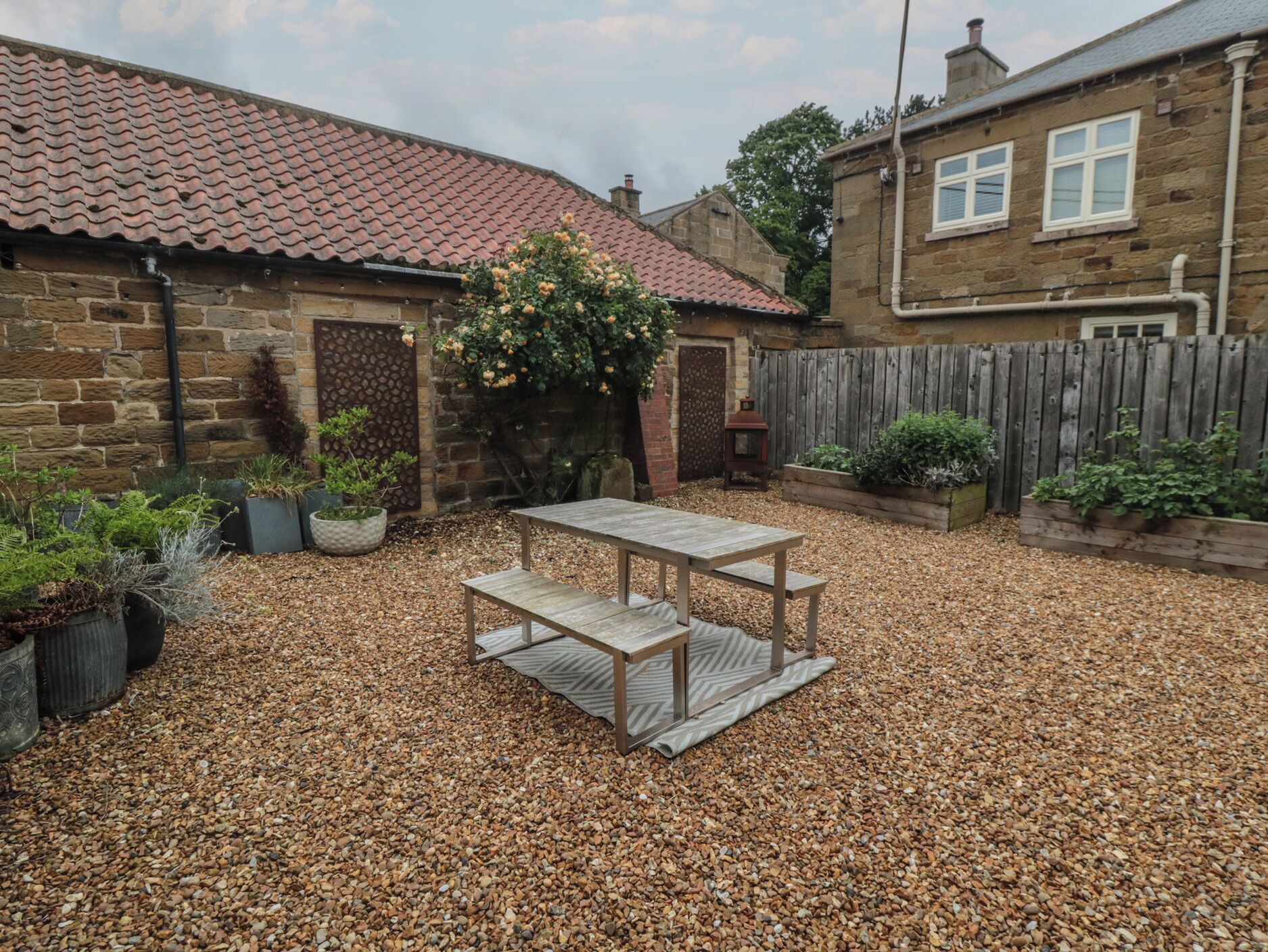 Un jardin avec une table et des bancs en bois, entouré de murs en pierre et d’une clôture en bois.