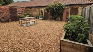 Un bâtiment en briques surmonté d’un toit en tuiles, avec une terrasse en gravier, une table en bois et des plantes en pot.