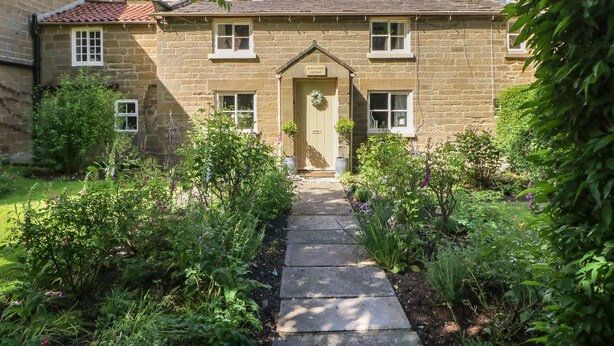 Une maison en pierre avec une porte centrale, entourée d’un jardin où poussent différentes plantes et fleurs.