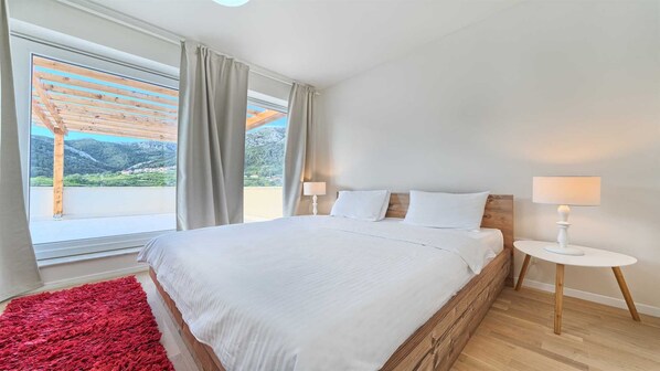 Un dormitorio con una cama grande, dos mesitas de noche, un tapete rojo y vistas a montañas desde la ventana.