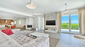A modern living room with a large grey sectional sofa, a flat-screen TV mounted on a stone wall, and a dining area with a wooden table and chairs. - Villa Luxoria - for 10 guests with 4 bedrooms, a private pool and a spa by MyWaycation (Jelsa)