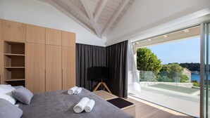 A modern bedroom with a large bed, a TV, and a balcony with a view of the sea.