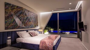 A modern bedroom with a large bed, a ceiling-mounted TV, and a large window overlooking a night scene.