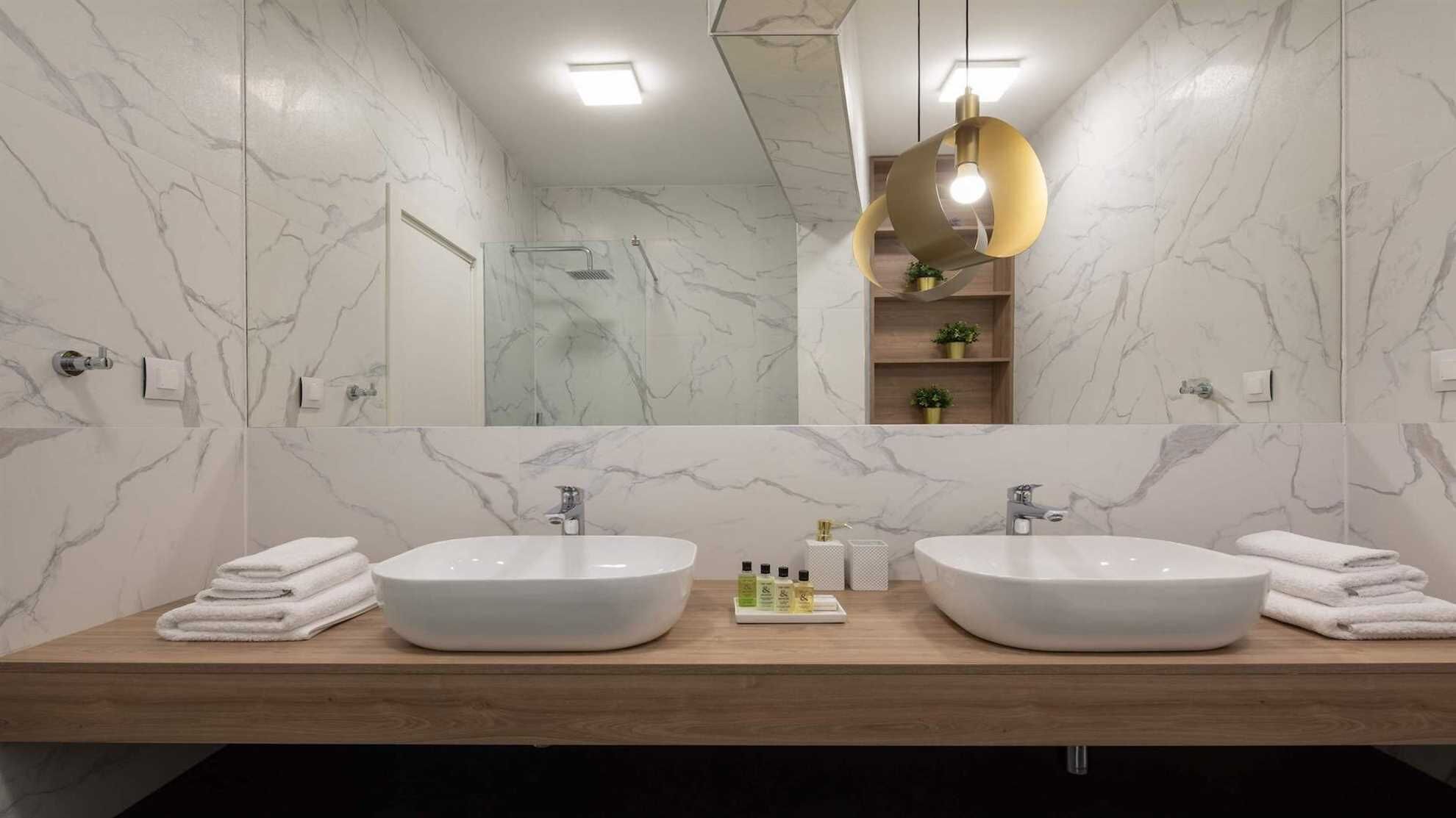 A bathroom with two white marble sinks, a wooden vanity, a large mirror, and a gold pendant light.
