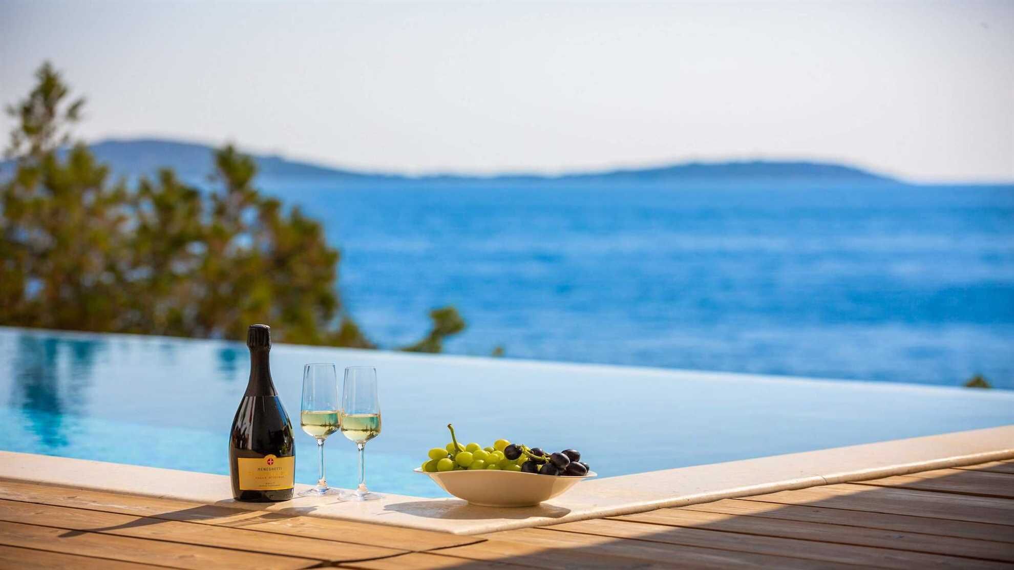 A wooden deck with a bottle of wine, two glasses, and a bowl of grapes by a pool with a view of the sea and mountains.
