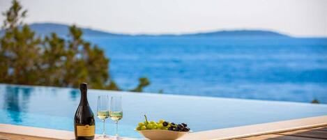 A wooden deck with a bottle of wine, two glasses, and a bowl of grapes by a pool with a view of the sea and mountains.