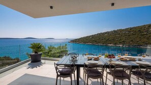Une table à manger dressée avec des assiettes, des verres et un plateau de fruits, offrant une vue sur la côte et un ciel dégagé.