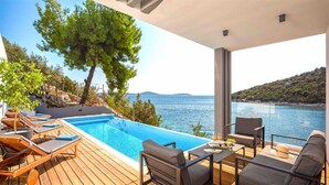 A poolside area with lounge chairs and a table, overlooking a body of water and a hilly landscape. - Villa Endless Summer - 5 Star beachfront villa with a pool by MyWaycation (Vinišće)