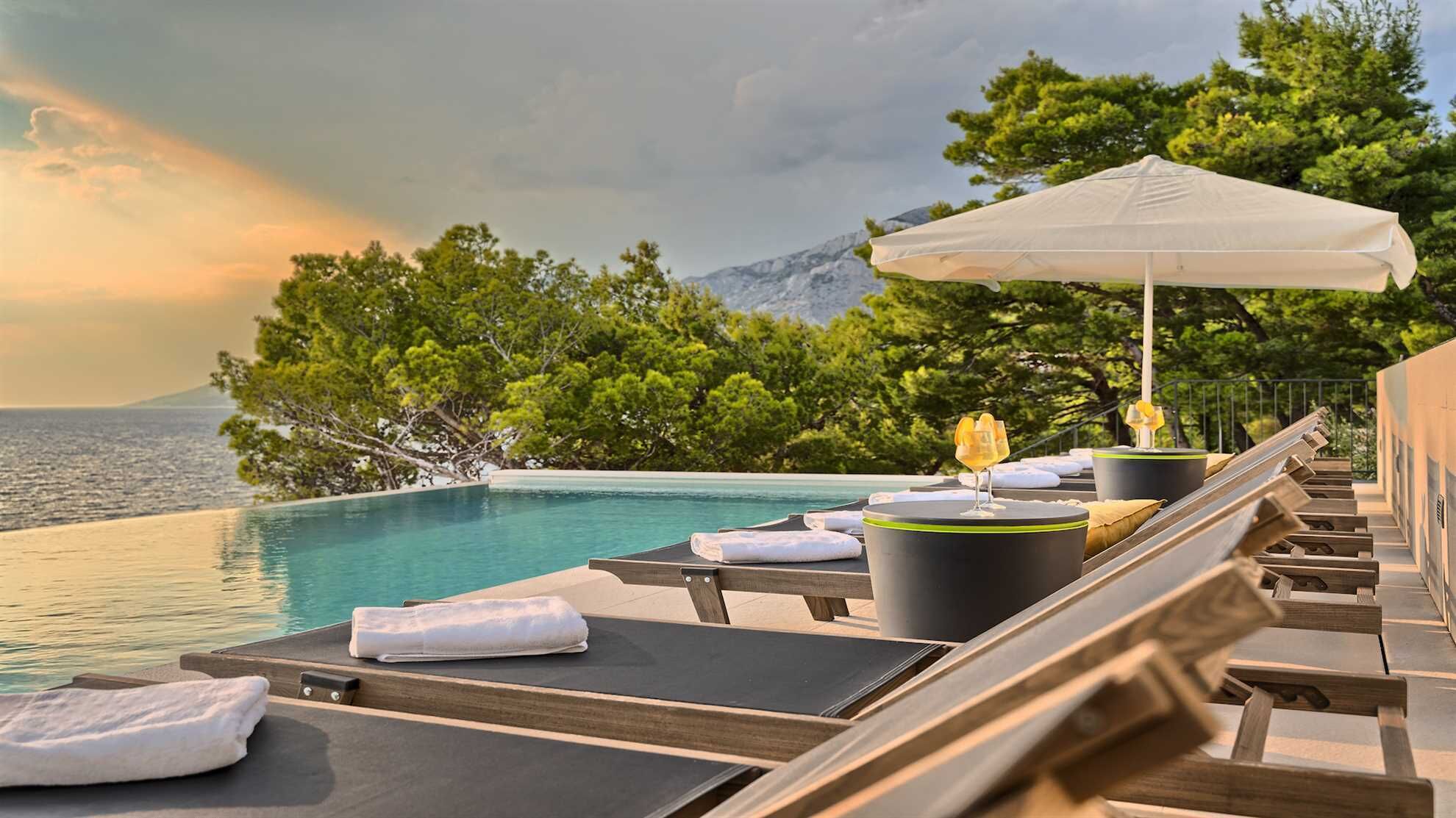 Uma área à beira da piscina com espreguiçadeiras, uma mesa com bebida e vista para o mar e árvores.