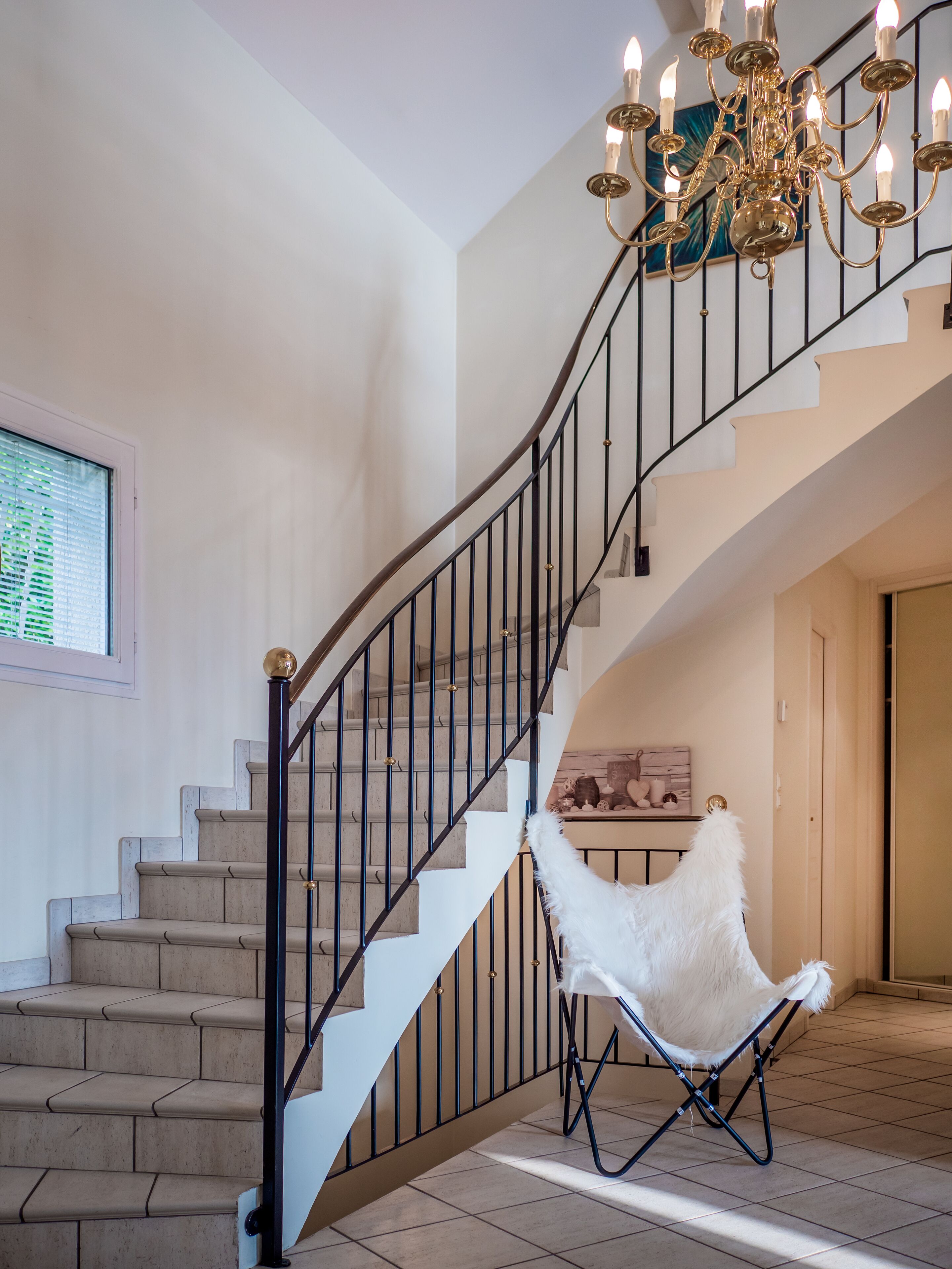 Una escalera con barandilla de metal negro, peldaños color crema y una butaca mullida de color blanco al pie.