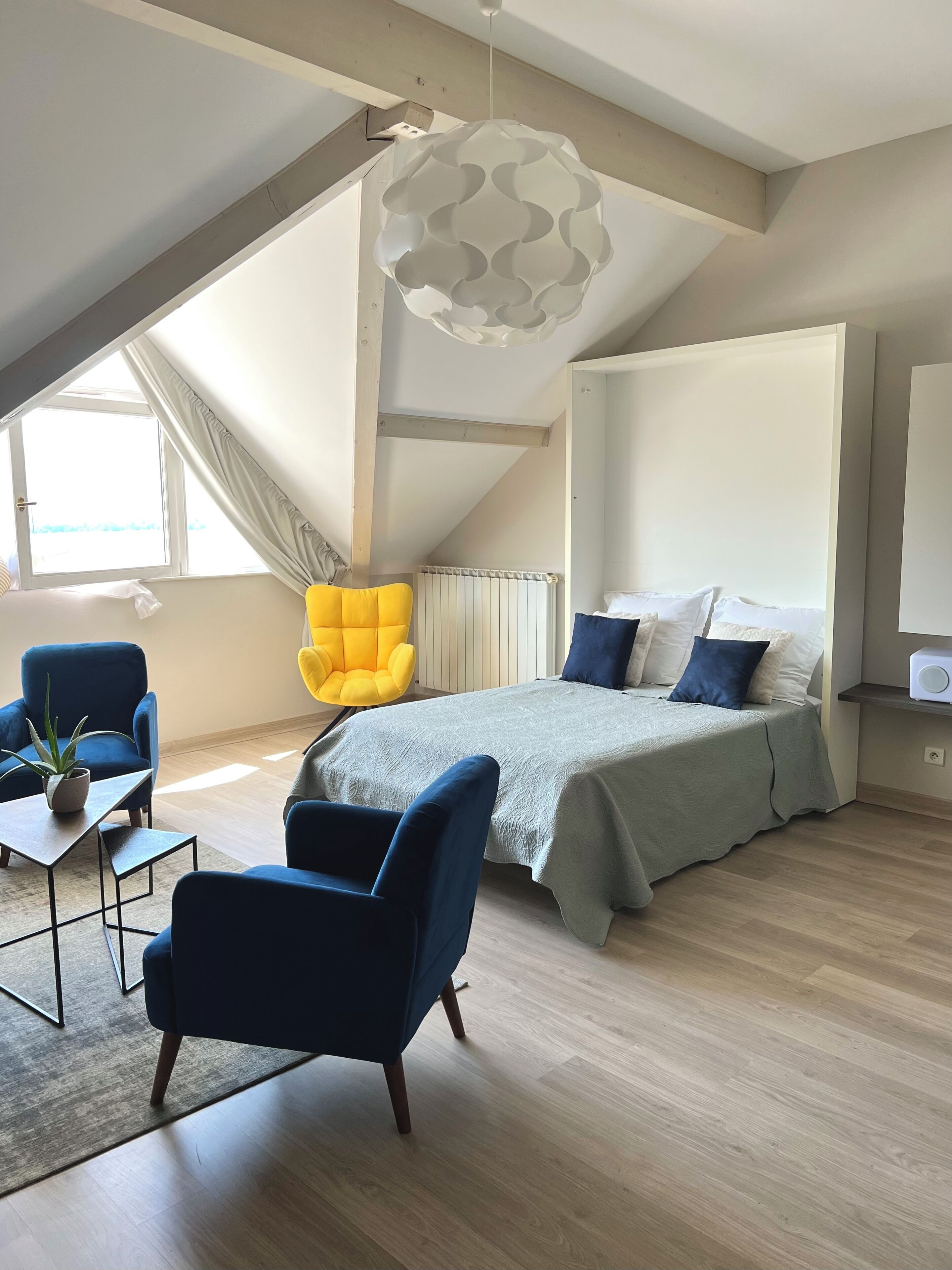 Een moderne slaapkamer met een schuine plafond, een bed met blauwe en witte beddengoed, een gele fauteuil en een bijzondere hanglamp.