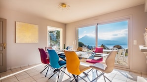 Un coin repas avec une table dressée pour six personnes, des chaises colorées, une grande fenêtre donnant sur les montagnes et un luminaire au plafond.