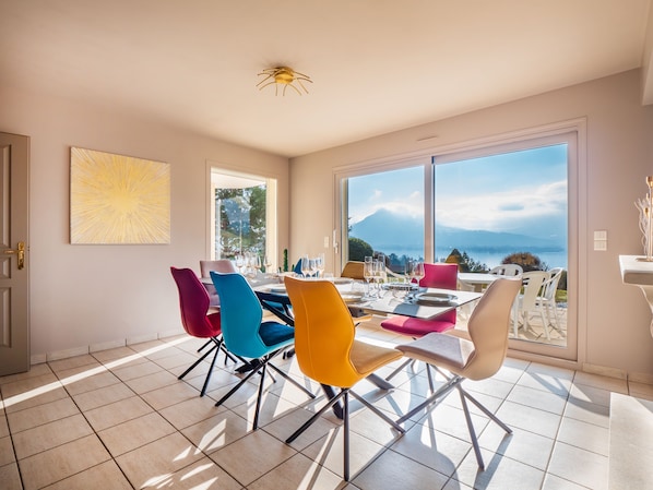 A dining area with a table set for six, colorful chairs, a large window with a mountain view, and a ceiling light fixture.