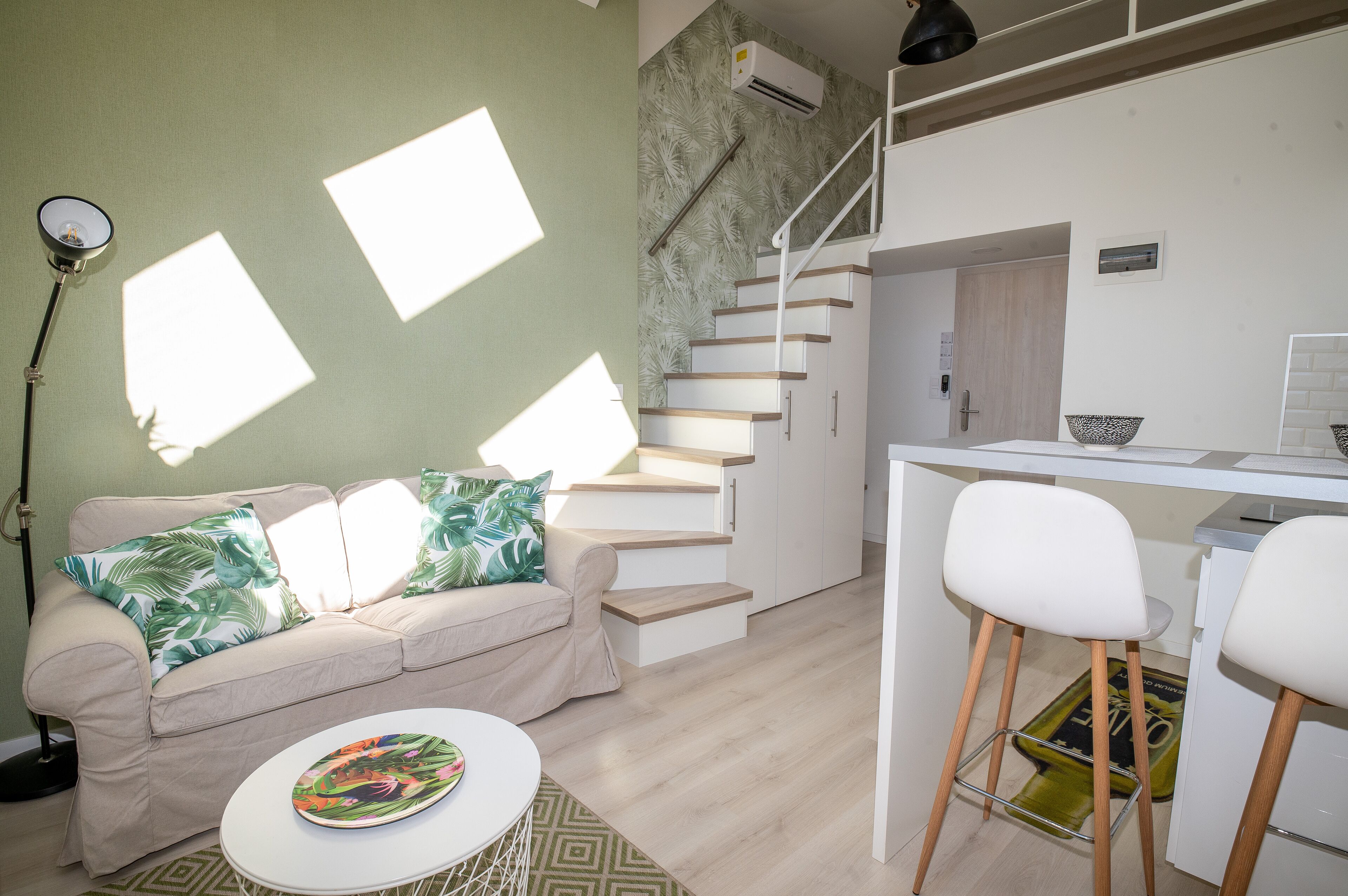 Ein moderner Wohnbereich mit Treppe, einer Couch mit tropischen Kissen, einem Essbereich mit Barhocker und einer Küchenzeile.